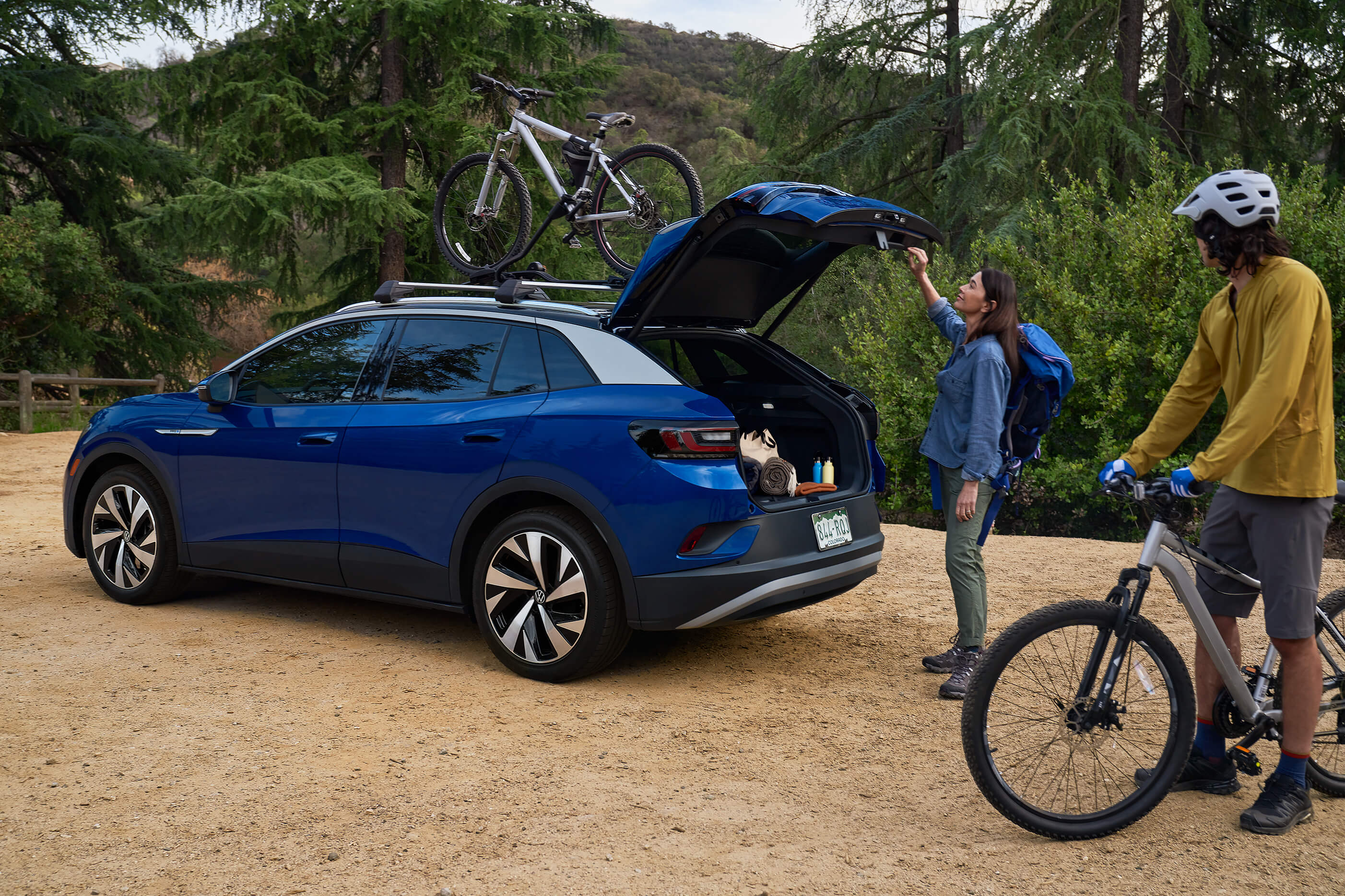A man and woman load mountain bicycles onto an ID.4 in Dusk Blue Metallic with cross bars and a bike attachment that's parked in a natural, outdoor setting.